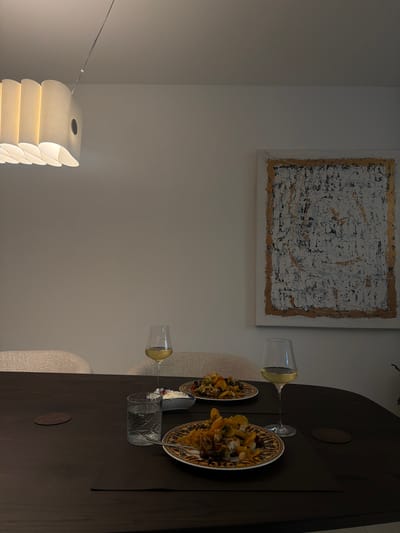 The image depicts a dining scene with two place settings on a dark wood table. Each setting includes a plate of food, a wine glass filled with white wine, and a small dish of what appears to be a creamy dip. A glass of water is also present at one setting. In the background, two chairs with upholstered seats are visible, and a large abstract painting hangs on the wall. A modern pendant light fixture hangs from the ceiling. The lighting in the room is dim, suggesting it might be evening. The scene appears to be set in a dining room or a well-lit area of a home. No people are visible in the image. The image was captured in Ridderkerk, Netherlands.