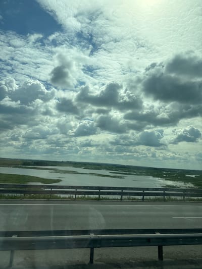 The image shows a wide, calm body of water with green reeds growing along its edges, likely part of the Kama River near Menzelinsk, Russia. In the foreground, a highway runs parallel to the water, separated by metal guardrails. The sky is filled with dramatic, volumetric clouds, some of which are dark and shadowed, suggesting a dynamic weather system. The sun appears to be shining through breaks in the clouds, casting a bright, diffused light. There are no people or vehicles visible in the image, giving the scene a sense of natural beauty and tranquility. The overall impression is of a vast, open landscape under a cloudy sky.