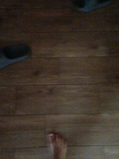 The image is an indoor shot of a wooden floor. In the bottom right quadrant, a human foot is visible, appearing to be a left foot. The foot is bare and angled towards the viewer. The rest of the image is dominated by the texture and grain of the wooden floorboards, which are laid horizontally. There is some dark shading in the upper left portion of the image, possibly indicating the presence of an object or shadow, but it is not clearly discernible. The lighting in the scene is dim, contributing to the grainy texture of the photograph. The scene does not provide any specific clues about its location within Novotitarovskaya, Russia, nor does it offer information about any particular actions or interactions beyond the presence of the foot on the floor.