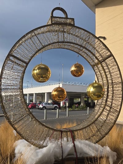The image depicts a large, circular, illuminated Christmas ornament structure situated outdoors in Blainville-sur-Orne, France. The primary subject is a dark metallic wire mesh frame, approximately 2.5 meters in diameter, with integrated small white LED lights and topped by a decorative cap and loop, designed to resemble a giant bauble. Four reflective, spherical, golden ornaments are suspended by thin lines within the central void of the circle. The base of the structure is adorned with dried, light brown ornamental grasses and white, synthetic wadding, simulating snow, with visible red securing straps. Through the center of the ornament, a partially cloudy blue sky is visible above a modern, multi-story building featuring white and light-toned facades. Below the building, a paved area, identified as a parking lot, contains several light-colored bollards and two identifiable vehicles: a red hatchback and a white SUV. The scene is under natural daylight, with the structure's integrated lights also active. No individuals or active interactions are visible.