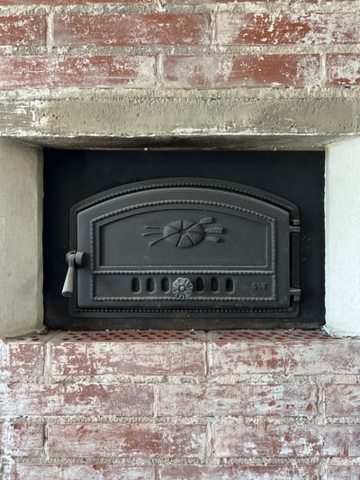 The image shows a close-up of a vintage cast-iron oven door set within a brick fireplace. The oven door is dark gray, ornately decorated with a central motif of wheat stalks and a sunburst, and features a handle on the left side and a decorative knob on the right. Below the central motif are several small oval vents and the letters "ST" are visible on the right. The fireplace opening is lined with black material, and the surrounding brickwork is red and aged, with white mortar showing signs of wear and peeling. The overall impression is one of rustic charm and historical character. This specific oven door is located in the historic Aulanko area of Hämeenlinna, Finland, likely part of a restored building or a historical attraction. There are no people or actions visible in the image, making it a still-life composition focusing on the details of the oven and its setting.