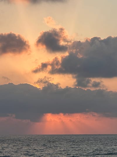 The image depicts a dramatic sunset over the sea, likely in the Mate Asher Regional Council, Israel, given the context. The sky is filled with a vibrant array of colors, from soft oranges and pinks to deep grays and purples in the clouds. Sunlight pierces through the cloud cover in distinct rays, creating a luminous effect. The sea below is a deep, textured gray, with small whitecaps visible. On the horizon, several distant, dark silhouettes suggest small boats or possibly distant landforms. There are no people or discernible structures visible in the immediate scene.