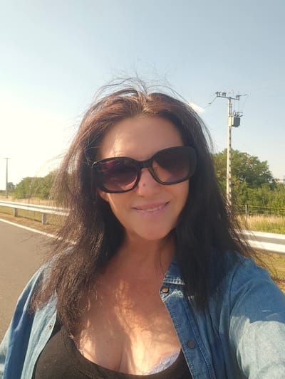 A middle-aged woman with dark hair and sunglasses is taking a selfie outdoors. She is wearing a denim shirt over a black top. In the background, a road with a white barrier runs alongside a field and trees. A utility pole with electrical equipment is visible in the distance. The sky is clear and blue. The location is likely Hidasnémeti, Hungary, as indicated by the presence of the distinctive utility poles and road infrastructure common in the region.