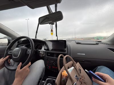 The image provides a first-person perspective from inside a car, an Audi, traveling on a road in Quart de Poblet, Spain. The weather is overcast and rainy, with numerous raindrops visible on the windshield and the car's wipers in an upright position.
The driver's hands are gripping the Audi steering wheel on the left side of the frame. On the right, a passenger's leg, clad in blue jeans, is visible, and their hand holds a blue electronic device, possibly an e-cigarette or vape. Resting beside the passenger's leg is a beige handbag, containing a bottle labeled "VapSolo" and a small card or paper showing the text "zavant."
The car's interior features a dashboard with a central infotainment screen displaying "13:36," indicating the afternoon, along with what appears to be a media or navigation interface. Below the screen, the climate control panel reads "Audi concert" and shows temperature settings of "21.0°" and "10°." An air freshener with a yellow liquid hangs from the rearview mirror. A tablet with a stylus rests on the dashboard above the glove compartment. An Audi logo is prominent on the steering wheel and subtly on the dashboard trim.
Outside, the road appears to be a multi-lane highway or main road, with streetlights stretching into the distance under the gray sky. Other vehicles are faintly visible through the rain. A green circular sticker with a prominent "C" is affixed to the lower right of the windshield, likely a Spanish environmental classification badge.
