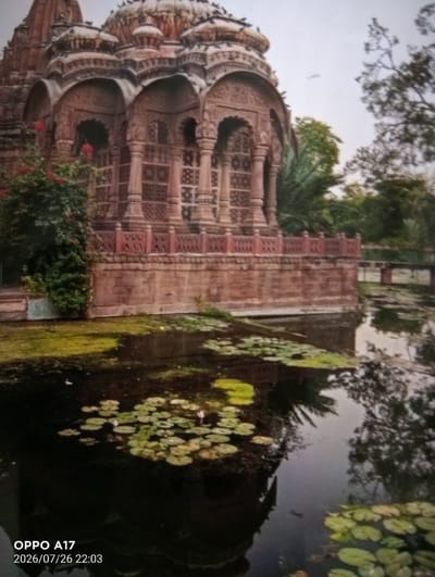The image depicts a temple-like structure with intricate carvings, constructed from reddish-brown stone. The structure has multiple arched openings and is surrounded by lush greenery. In the foreground, a body of dark water is visible, covered with lily pads and other aquatic vegetation. The water reflects the surrounding trees and the sky, which appears overcast. A bridge can be seen in the background, spanning the water. The overall scene is serene and appears to be a religious or historical site. The timestamp in the lower left corner indicates the image was taken on July 26, 2026, at 22:03, and was captured by an OPPO A17 device. The location is specified as Құмөзек, Kazakhstan.