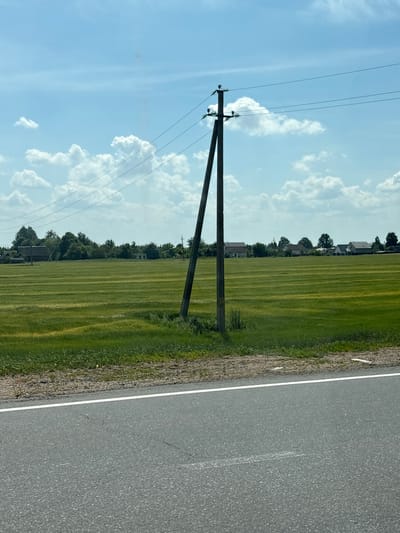 This media file depicts a rural roadside scene in the Мастоцкі сельскі Савет, Belarus. The image is oriented vertically, with the sky and a field of green grass dominating the left side, and a paved road occupying the right side.
In the foreground on the left, a wooden utility pole with multiple wires stretches upwards into a bright blue sky dotted with fluffy white clouds. The field is characterized by distinct rows, suggesting it is a cultivated crop, possibly wheat or barley, due to its yellowish-green hue. In the middle ground, several houses with roofs are visible amidst a line of trees. The road on the right is asphalt, with a white line marking its edge. The overall impression is a bright, sunny day, likely during daytime given the clear sky and sunlight illuminating the landscape. There is no visible text in the image. The scene conveys a sense of tranquility and rural simplicity.
