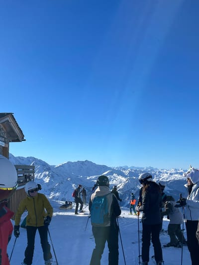 The image captures a vibrant winter scene in the snowy mountains of Séez, France, under a clear, bright blue sky. The shot is composed from a slightly elevated perspective, looking out over vast snow-covered peaks that stretch across the horizon.
Several people, presumably skiers, are gathered in the foreground and mid-ground of the image, equipped with ski gear including helmets, goggles, skis, and poles. In the immediate foreground-right, a person in a bright yellow jacket and black pants stands on skis, wearing a white helmet. To their left, another person in a white helmet and red jacket is partially visible. Further back, a skier with a light blue backpack and a black helmet stands, while another individual wears a dark jacket and a white helmet. Towards the top-right, a person stands out with a distinctive white, fluffy unicorn-themed helmet cover. All individuals appear to be taking a break or waiting on a ski slope, facing the expansive mountain range.
The weather is bright and sunny, with strong sunlight casting clear shadows. Visible sun rays pierce through the upper left portion of the sky, indicating a strong light source. The only clouds are very faint, high-altitude wisps.
In the foreground-left, a rustic wooden structure, possibly a signpost or part of a small hut, is visible. Text on this structure includes "EN COURS" (in progress) and "PISTE VERTE" (green slope), suggesting they are on an easy ski run. Another wooden barrier or fence is also present near the foreground skiers. The overall atmosphere conveys a cheerful day for winter sports amidst a breathtaking alpine landscape.