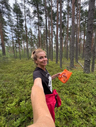 A woman, with blonde hair pulled into a bun and wearing hoop earrings, is taking a selfie in a forest. She is wearing a black t-shirt and a red jacket tied around her waist. She is holding an orange berry-picking comb in her right hand. The forest is filled with tall pine trees and lush green undergrowth, characteristic of the Kliuchevoe region in Russia, which is known for its forests and berry-picking. The sky is overcast, suggesting a cloudy day.