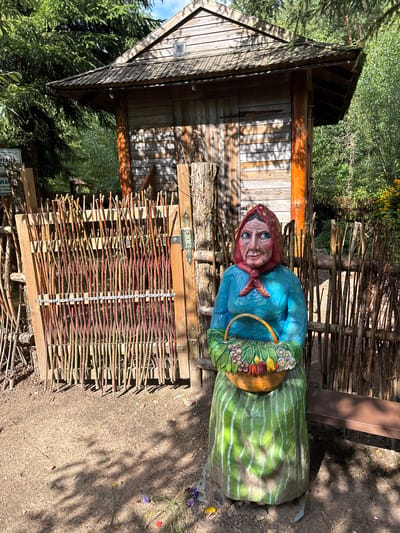 The image depicts a carved wooden statue of an elderly woman sitting in front of a rustic wooden shed in Talkowszczyzna, Poland. The woman wears a blue blouse, a red headscarf tied with a knot, and a green and white striped skirt. She holds a woven basket filled with flowers and vegetables. The shed is constructed from rough-hewn wooden planks and has a shingled roof. A fence made of intertwined branches encloses the area. The ground is covered in gravel and dirt, with scattered wildflowers near the statue's base. Sunlight casts dappled shadows across the scene. A brown park bench is visible to the right of the statue.