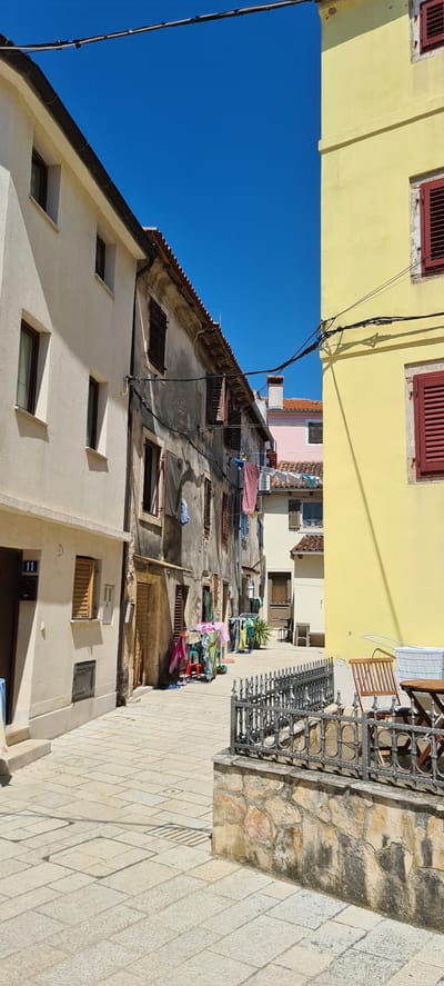 This is an outdoor, eye-level, medium shot of an alleyway with buildings on both sides, taken on a clear, sunny day. The scene depicts an urban environment, possibly in Općina Fažana, Croatia, characterized by narrow streets and older architecture.
On the left, a bright yellow building occupies the upper part of the frame, featuring two windows with red shutters. Below this, a beige building with a sloping roof and several windows is visible. The upper portion of this building shows a tiled roof and some laundry hanging on clotheslines strung between buildings. Further down the alley, more buildings are visible, some with peeling paint and weathered facades, suggesting age and history. Various objects are scattered around, including potted plants, chairs, and more laundry.
On the right side of the alley, a walkway paved with light-colored stones leads into the distance. A decorative wrought-iron railing runs along a stone wall, separating the walkway from the buildings. Several metal grates are set into the stone pavement. A number "11" is visible on a dark blue sign above a doorway on the beige building.
The bright blue sky suggests it is daytime, likely mid-morning or afternoon given the strong shadows cast by the buildings. The overall impression is one of a Mediterranean town, with its characteristic narrow streets, colorful buildings, and everyday life unfolding outdoors. There are no people visible in the frame, but the presence of laundry and outdoor furniture implies recent or current human activity.