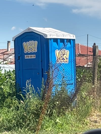 The image shows a portable toilet with "Yes!" branding and a phone number "0700 500 50" displayed on its side. The toilet is blue and white and is situated outdoors, surrounded by overgrown green vegetation and a chain-link fence. In the background, there are residential buildings with tiled roofs and chimneys, indicating an urban or suburban environment. The sky is clear and blue with a few small white clouds and two distant flying objects, possibly birds or drones. The scene appears to be in the city of Petrovo, Bulgaria, suggested by the provided context. There are no people or actions depicted in the image.
