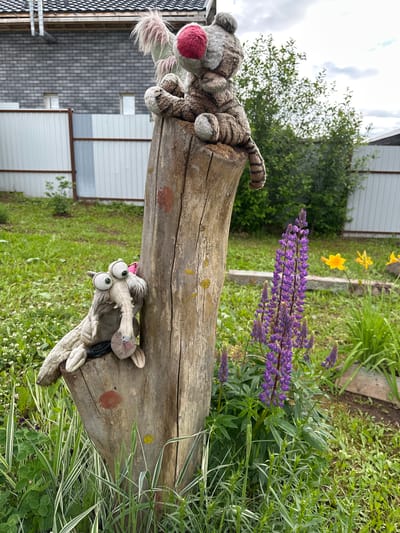 An outdoor scene in Bol'shaya Mos', Russia, depicts two plush toys positioned on a weathered wooden tree stump. A striped, tiger-like plush with a prominent red nose and a pinkish fur tuft on its head is seated on the top of the stump. Below it, leaning against the stump, is a grey, long-snouted plush toy with large, spherical eyes, which holds a small, furry, light-brown object. The stump itself is vertically cut, exhibiting rough bark texture, and has a red circular mark and a smaller yellow mark painted on its surface.

The ground surrounding the stump is covered in green grass, interspersed with small white flowers and variegated green and white plant foliage in the foreground. To the right of the stump, a cluster of vibrant purple lupine flowers stands tall, with yellow daylilies visible in the background. Dense green foliage from bushes and trees forms a middle ground layer, partially obscuring a corrugated metal fence, which is white and grey with brown support posts. In the far background, a grey brick building with a dark tiled roof and exposed utility pipes is visible, featuring two small rectangular windows. The sky is overcast with diffused cloud cover.