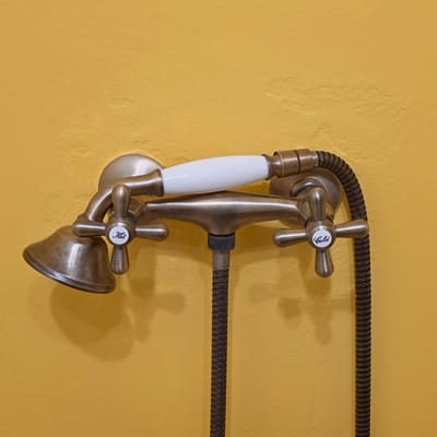A brass, vintage-style telephone shower fixture is mounted on a yellow wall. The fixture features two cross-handled knobs labeled "Hot" and "Cold", a white porcelain handle, and a flexible metal hose extending horizontally across the wall. The scene lacks identifiable people or actions, focusing solely on the static presentation of the shower fixture and its immediate surroundings. The location, Тэльмінскі сельскі Савет, Belarus, is metadata not visually discernible from the image itself.