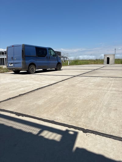 A dark blue van is parked on a concrete surface. The van is viewed from the rear passenger side, and its rear doors are closed. The van has tinted windows and black trim around the wheel wells and along the lower body. The license plate, visible on the rear of the van, reads "BJ 932DC".

The concrete surface appears to be a large, open area, possibly an industrial or training ground, with expansion joints running across it. In the background, a tall concrete structure with a dark opening is visible. To the left of this structure, a grassy area with some foliage is present. Several sprinklers are active in the grassy area, creating a misty effect. A metal structure with stairs and railings is also visible to the left of the van, partially obscured by the van.

The sky is a clear, bright blue, indicating a sunny day. The angle of the shadows cast on the concrete suggests it is late morning or early afternoon. The overall scene appears to be a functional outdoor area, possibly related to training or operations, given the presence of the concrete structure and sprinklers. The image is taken in Pivdennoukrainsk, Ukraine.
