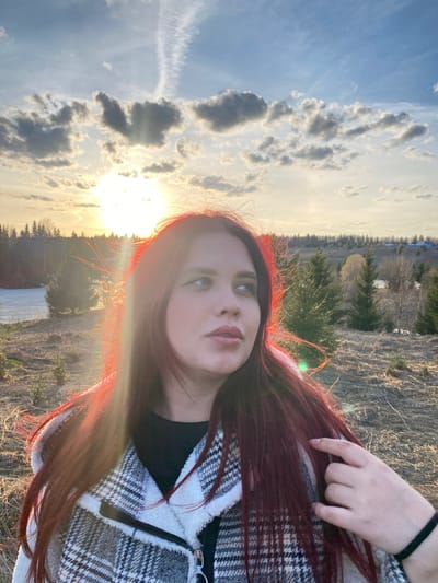 A young woman with long auburn hair is posing for a selfie in a field. She is wearing a black shirt and a plaid, sherpa-lined jacket. The location appears to be in the area of Ореховая Гора, Russia, based on the landscape visible in the background, which includes fields, trees, and distant structures. The sky is visible with clouds and the sun setting. The sun is causing a lens flare around her hair. One of her hands is visible touching her hair.