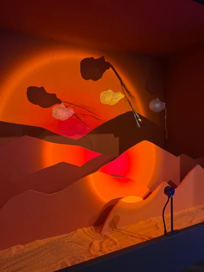 An artistic diorama in Sélestat, France, depicts a stylized desert or alien landscape under artificial sunset illumination. The scene is contained within a dark-framed enclosure. The base layer consists of fine-grained, orange-red material resembling sand. Layered, undulating cutouts, colored in muted orange and reddish-brown, form a mountainous or dune-like topography in the midground and background.

A circular, intense orange light projects onto the upper rear wall, simulating a setting sun. Below this, another smaller, circular red light illuminates a section of the middle ground. Four translucent, sculpted objects, resembling jellyfish or bioluminescent flora, are suspended from the top of the enclosure by thin lines. The leftmost object is white, multi-lobed, with dark, wire-like tendrils extending downwards, casting a prominent, elongated shadow onto the back wall. Two central objects include a yellowish, single-lobed form and another white, multi-lobed form with tendrils, both illuminated by warm light and casting shadows. The rightmost suspended object is a white, single-lobed form. Some of these objects appear to be attached to flexible stems containing small embedded lights, creating glowing points.

On the front right, a black, cylindrical projector device with a blue band is mounted vertically on a dark base, pointing into the scene, likely responsible for some of the projected light effects. The overall lighting creates strong contrasts and elongated shadows, enhancing the depth and otherworldly aesthetic. No people are present.