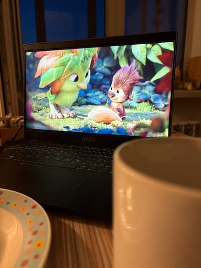 The image shows a close-up view of a laptop screen displaying a colorful animated scene with two characters. One character, on the left, is a green creature with large eyes and vibrant, feathered adornments. The other character, on the right, is a furry, brown creature with wide eyes and a somewhat curious expression. They are set against a lush, green, and blue background with floral elements.
To the right of the laptop, a portion of a white mug is visible, obscuring part of the screen. Below the laptop, a small plate with a floral pattern is partially visible. The laptop keyboard is black and has Cyrillic lettering on the keys, suggesting it is set to a Russian language layout. The overall scene appears to be indoors, possibly in a home environment, given the presence of the laptop, mug, and plate. The lighting is dim, and the image is taken at an angle, with the screen dominating the left side of the frame. There is no indication of the time of day or weather conditions. The emotion conveyed by the animated characters is one of engagement and possibly playfulness. The text "IIISII" is visible on the laptop's bezel.