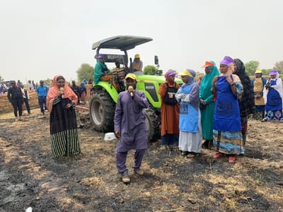 A group of people are gathered in an open field, likely in Ringimawa, Nigeria. The field appears to have recently been burned as the ground is charred and blackened. In the center of the gathering, a woman in a black and green dress with a head scarf holds a microphone and speaks to the group. Behind her, a green tractor sits facing away from the group. There are multiple men and women in the group, some of whom are wearing blue aprons with the phrase "NGAN LIFE" in a circular design, while others wear similar blue aprons with the phrase "NGAN BARDA," possibly related to an agricultural initiative or community project. The environment is dry and dusty.