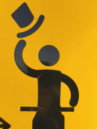 The media file is a close-up, eye-level shot of a black silhouette icon on a bright yellow background. The silhouette depicts a person wearing a top hat, which they are tipping or doffing. The figure has a circular head, a torso, and one arm raised in a gesture of greeting or acknowledgment, holding the hat. The lower part of the body is cut off by the bottom of the frame, but appears to be standing on a rectangular platform. The style of the icon is simple and graphic, suggestive of a public sign or pictogram. The yellow background is textured, possibly indicating a painted surface or a sticker. There are no discernible cues for time of day, weather, or specific emotions, other than the polite gesture conveyed by the icon. No text is visible within the frame. The location context is Tczew, Poland.