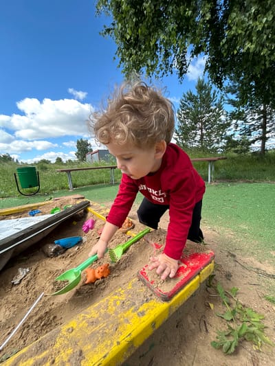 The image depicts a young child playing in a sandbox. The child, with curly blonde hair, is wearing a red long-sleeved shirt and black pants. The sandbox is filled with sand and contains various toys, including a green shovel and an orange pig-shaped toy. The child is actively engaged in playing with the sand and toys. The background shows a grassy area, trees, a bench, and a green bin, suggesting an outdoor public space. The sky is blue with some clouds. There is no specific information in the image to indicate the location is the city of Борщевка Малая, Russia.