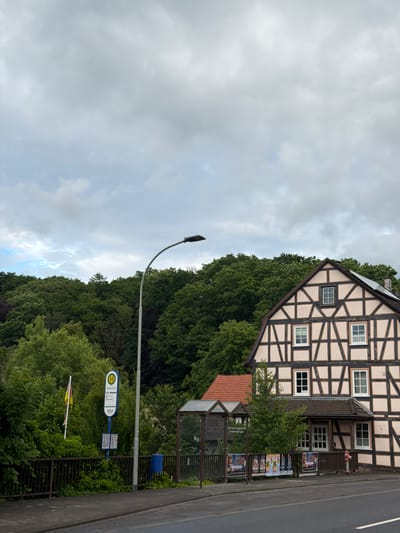 The image depicts a street scene in Kirtorf, Germany. On the left, a bus stop sign with the "H" symbol is visible, indicating a public transportation stop. A flag with yellow and red stripes is mounted on a pole nearby. Adjacent to the bus stop, there is a bus shelter. In the center and right of the image, a half-timbered building with light pink walls and dark brown beams is prominent. This building has several windows and a tiled roof. In front of the building, a row of posters is displayed, and a red fire hydrant stands next to the entrance. The street is paved, and a sidewalk runs alongside it. A street lamp arches over the scene. The background is dominated by a dense forest of green trees under a cloudy sky.