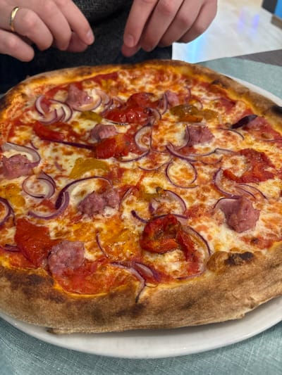 A close-up, high-angle shot depicts a freshly baked pizza served on a white round plate, resting on a light blue textured tablecloth. The pizza features a golden-brown, well-risen crust with localized charring. Its surface is covered with a melted white cheese and tomato sauce base, topped with numerous slices of red onion, irregular chunks of cooked sausage, roasted red pepper pieces, and some scattered yellow pepper segments. In the upper background, the hands of an individual are visible, positioned above the pizza. The left hand displays a gold band on the ring finger. The individual is wearing a dark, possibly grey, long-sleeved garment. The scene takes place indoors, likely within a restaurant or pizzeria in Ferrara, Italy.