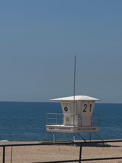 The image captures a lifeguard tower on a beach, most likely in Sant Pol De Mar, Spain. The tower, marked with the number "21", has a white body with the inscription "112" and decorative arrow motifs. It stands on stilts, with a ladder providing access. The sky is a clear, pale blue. The sea is visible beyond the tower, appearing calm. A portion of the sandy beach is visible to the right. A long, thin metal pole extends horizontally from the tower. The overall scene is bathed in bright daylight.
