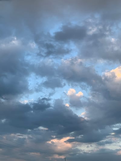 The image displays a sky view over Bratkivtsi, Ukraine, dominated by a complex formation of clouds. Dark grey, medium grey, and white clouds are layered and interspersed throughout the frame, with numerous breaks revealing segments of a lighter blue sky, particularly in the upper and central portions. Towards the bottom right and central areas, some clouds exhibit a soft pinkish-orange hue, suggesting indirect light illumination. Faintly visible in the lower left quadrant are several thin, horizontal dark lines consistent with overhead utility wires, with a partial view of a vertical support structure, likely a utility pole, at the very bottom center. No individuals, distinct ground-level objects beyond the utility infrastructure, or specific actions/interactions are present.