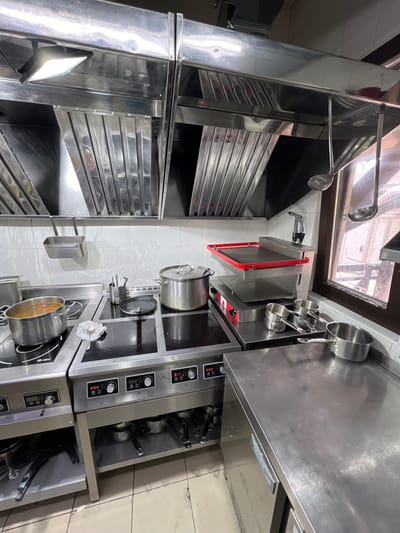 The image shows a commercial kitchen environment, likely in Plast univska, Russia. The scene is dominated by stainless steel surfaces and appliances. In the foreground, a stainless steel workbench is visible, with a handle on the left side. Behind it, a multi-burner induction cooktop is present, with various pots and utensils on its surface. One pot contains a liquid with some solid ingredients. To the right of the cooktop, a tiered cooking apparatus with a red-rimmed top shelf is set up. Further to the right, a window offers a view of an outdoor area with wooden structures. Above the cooking area, a stainless steel exhaust hood is installed, with ladles hanging from it. The walls are tiled with white square tiles. The overall impression is of a functional, busy kitchen, though no people are visible in the frame.