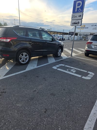 A black SUV is parked in a parking lot, its tires on a white-lined, handicapped parking space. The space is marked with a white wheelchair symbol painted on the asphalt. A signpost stands next to the parking spot, displaying three signs. The top sign is blue with a white wheelchair symbol and a white "P". Below it is a rectangular sign with a white arrow pointing to the right and the text "25m". The bottom sign is a white rectangle with a silhouette of a car and a white arrow pointing to the right. In the background, a modern building with large windows and some retail signage is visible. The building has "РЕМАНа" and "СА" written on it in large letters. Other cars are parked in the vicinity, and a person is visible near one of them. The sky is partly cloudy with a bright, hazy sun, suggesting it is either early morning or late afternoon. The scene appears to be a public parking area, possibly outside a shopping center or commercial establishment.