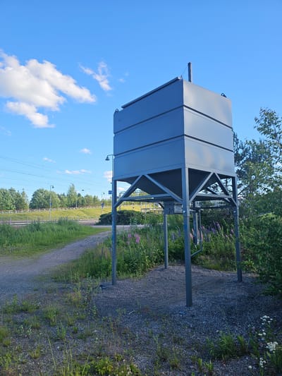 The image, taken in Mäntsälä, Finland, depicts a large, metallic, rectangular structure supported by four grey metal posts. The structure has a sloped roof and appears to be elevated above the ground. The metallic surface is smooth and reflects the bright blue sky with scattered white clouds. Green vegetation, including grass and flowering plants with purple and white blooms, surrounds the base of the structure and extends into the background. A gravel path runs alongside the vegetation. In the distance, there are power lines and more trees, suggesting an outdoor, possibly rural or semi-industrial setting. The lighting suggests it is daytime, with clear skies. There is some blue graffiti visible on one of the posts. The overall scene appears to be peaceful and serene.