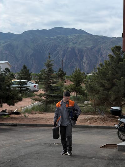 A young man walks on a paved road, carrying a black briefcase and wearing a grey work jacket with orange accents and a headset. He is dressed in dark pants and sneakers. In the background, a white car is parked next to a building. A mountain range with green, rugged slopes rises behind the scene, under a cloudy sky. The area appears to be outdoors, possibly in a semi-rural or suburban setting within Kaji-Say, Kyrgyzstan, given the presence of mountains and vegetation. A motorcycle is partially visible to the right.