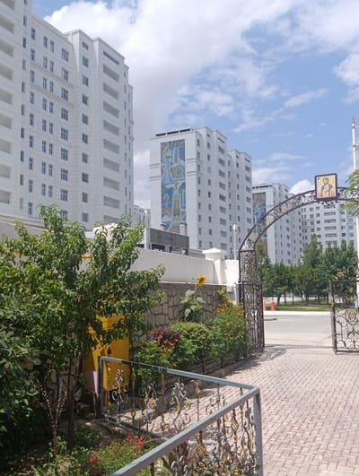 The image depicts a street scene in Ashgabat, Turkmenistan, on a sunny day with scattered clouds. Dominating the background are several tall, white, modern apartment buildings with numerous windows and balconies. These buildings are adorned with large, colorful mosaic murals. In the foreground, a decorative wrought-iron archway frames a path leading into a courtyard or garden area. The archway has ornate scrollwork and is topped with a framed icon of a religious figure. To the left of the archway, lush green trees and bushes are visible, along with a stone wall and a bright yellow structure. A single sunflower stands near the wall. The ground in the foreground is paved with light-colored bricks arranged in a herringbone pattern, and there is a metal railing along the path. The overall impression is one of urban development with artistic embellishments.