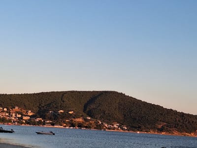 A wide shot captures a tranquil seaside scene in Erdek, Turkey, under a clear blue sky. The sun appears to be low on the horizon, casting a warm glow that softly illuminates a lush, tree-covered hillside dotted with white buildings. This hillside slopes down towards a calm, blue body of water.

Several boats are visible on the water. In the foreground, a dark-colored boat is partially submerged at the edge of the frame. Further out, a sleek, white motorboat is moored. A series of buoys or markers are strung across the water, creating a subtle line. The water's surface reflects the soft light, creating a peaceful atmosphere.

The hillside is densely covered in green foliage, with clusters of white and cream-colored houses nestled among the trees and along the coastline. The architecture suggests a Mediterranean or coastal style, with many of the homes featuring flat roofs and simple designs. The overall impression is one of a quiet, sun-drenched coastal town.

The time of day appears to be either early morning or late afternoon, given the angle of the light and the softness of the shadows cast on the hillside. The weather is clear and calm, with no indication of wind or rough seas. The scene evokes a sense of peace and relaxation, suggesting a leisurely day by the sea. No text is visible in the image.