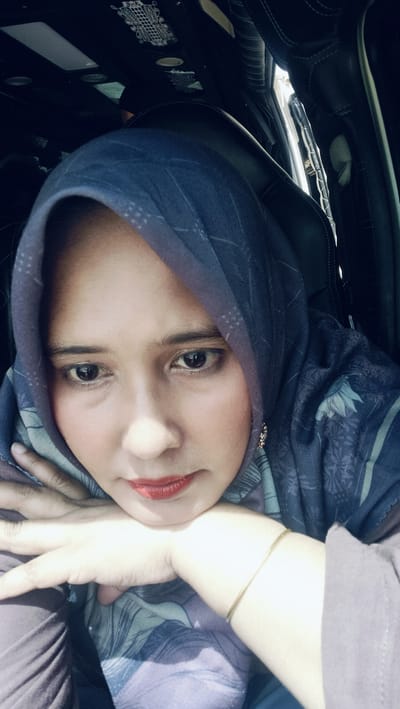 A selfie is taken from a low angle, showing a woman wearing a dark blue hijab with a patterned design. Her head is tilted, and her chin rests on her folded hands, which are placed on her chest. She is wearing subtle makeup, including red lipstick and eyeliner. A thin gold bracelet is visible on her left wrist. The background suggests she is inside a vehicle, with black car seats and interior details visible. The lighting appears to be natural daylight. The image is sharp and in focus on the woman's face. There is no identifiable information to link the image to Kutalawah, Indonesia, or any specific city.