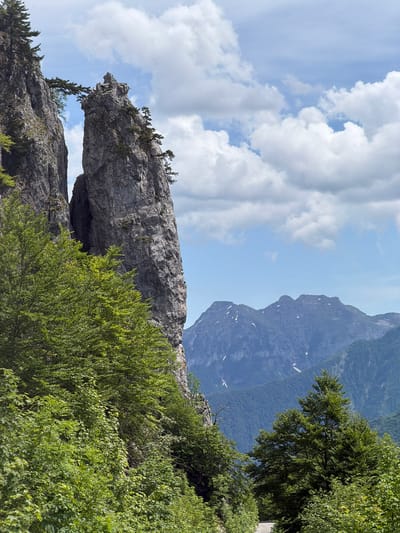 The image depicts a mountainous landscape in Dragovića Polje, Montenegro, under a partially cloudy sky. Two prominent, weathered grey rock formations dominate the left-center of the frame. The formation on the left is a sheer cliff face, while the one to its right is a tall, slender rock pillar or spire. A natural rock arch, formed by a single horizontal rock span, connects the upper sections of these two formations. Sparse green conifer trees and bushes cling to the ledges and summits of both rock structures and the arch.
In the foreground and midground, lush green deciduous trees and bushes cover the lower slopes and obscure the base of the rock formations. A narrow, winding paved road is visible in the lower right quadrant, bordered by dense green foliage, and appears to ascend into the distance. In the far background, hazy blue-grey mountains with scattered lighter patches, possibly snow or bare rock, rise against a lighter blue sky. The sky above is characterized by white cumulus clouds, more concentrated in the upper left and right, with clear blue sky visible elsewhere. No human figures or animals are present. The scene is illuminated by daylight, casting soft shadows.