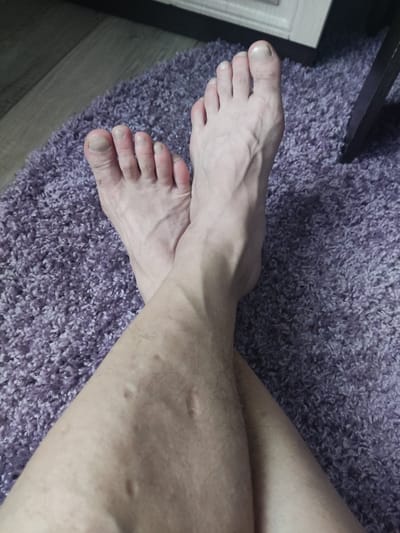 The image shows a close-up view of two crossed human feet, resting on a soft, purple, shag carpet. The feet are pale in skin tone, with visible veins on the top of the feet and ankles. The toenails are visible and appear unpainted. The individual's legs are also visible, showing some minor skin imperfections and hair. The background consists of the purple carpet and a sliver of what appears to be wooden flooring and furniture. This image does not contain any information that would allow for the determination of its location in Gorno-Altaysk, Russia.