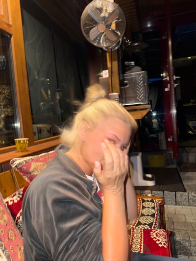 A medium shot captures a blonde woman in the foreground, her face partially obscured by her hands. She is seated on a bench with a richly patterned cushion. She wears a dark grey velour jacket over a white shirt. Behind her, a large window reflects a dark, indistinct interior. Above her head, a metal fan is mounted to the ceiling. To the right, a red-framed doorway leads to an outdoor area with stone steps. A yellow bucket sits on the ground near the doorway. The scene appears to be an indoor seating area, possibly a cafe or restaurant, with elements suggesting it is located in Heiligenrode, Germany, though specific landmarks are not visible.