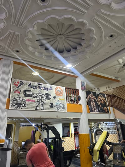 The image shows the interior of a gym located in El Hajeb, Morocco. The ceiling features ornate white plasterwork with recessed lighting and a central decorative element. A large mural on the wall depicts muscular men lifting weights and the text "The GYM" and "fitness". To the right of the mural are framed pictures of bodybuilders and trophies on display. The gym is equipped with various weightlifting machines and exercise equipment. In the foreground, a person wearing a pink t-shirt is partially visible, facing away from the camera and towards a weight machine. Another person in a white t-shirt can be seen in the background operating equipment. The overall environment suggests a functional, albeit somewhat dated, fitness facility.