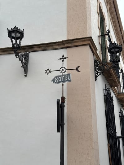 An ornate metal hotel sign is affixed to a pole at the exterior corner of a multi-story building in Ronda, Spain. The building features textured white stucco walls with a vertical beige-textured corner column and a horizontal brown trim line. The sign itself is crafted from dark metal, forming a directional arrow with the word "HOTEL" in white capital letters on a dark blue background within its shaft, pointing left. Above the "HOTEL" sign, the metalwork extends horizontally, incorporating a Star of David on the left, a central spoked wheel, and a crescent moon on the right. A cross surmounts the central wheel. Two ornate, dark wrought iron street lamps with decorative scrollwork brackets are mounted on the building's walls; one is prominently displayed on the left wall, and another is partially visible on the right wall, positioned above a window with dark green shutters and black wrought iron security grilles. A portion of an upper-level balcony with similar black grilles is visible on the right. No individuals are present in the scene.