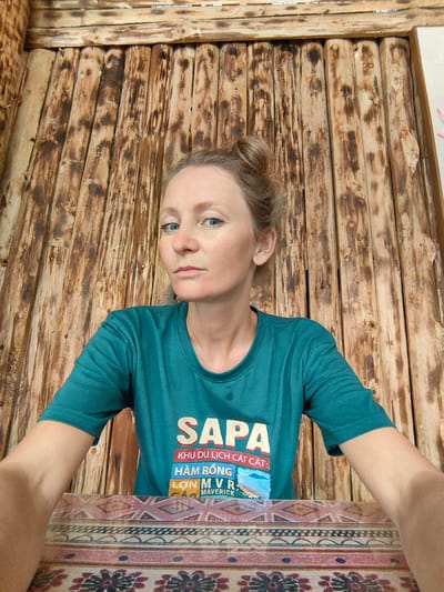 A medium shot captures a fair-skinned woman with blonde hair styled in a bun. She is positioned indoors, wearing a teal t-shirt with "SAPA" printed in large white letters above smaller text in Vietnamese and English, including "KHU DU LỊCH CÁT CÁT," "HÀM RỒNG," "LỚN MVR," and "MAVERICK." The background consists of vertical wooden planks with a rustic appearance. The location is identified as Thành phố Hà Giang, Vietnam.