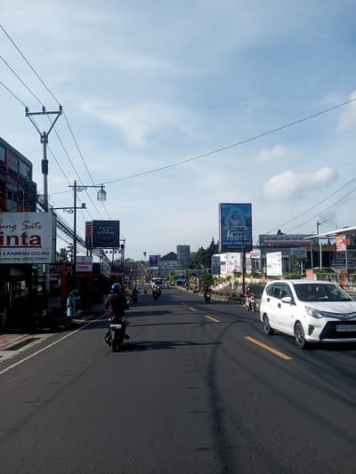 The image shows a street in what is likely a Southeast Asian city. A white car is driving on the right side of the road. Several motorbikes are also on the road. Utility poles with electrical wires are visible on the left side of the road. There are various signs and advertisements along the side of the road, including a sign that says "CHINESE FOOD" and "STURGIS." The sky is blue with some clouds. The license plate on the white car says "B 2240 BZH." The location cannot be determined to be Phou Khoun, Laos, as the signs are not in Lao.