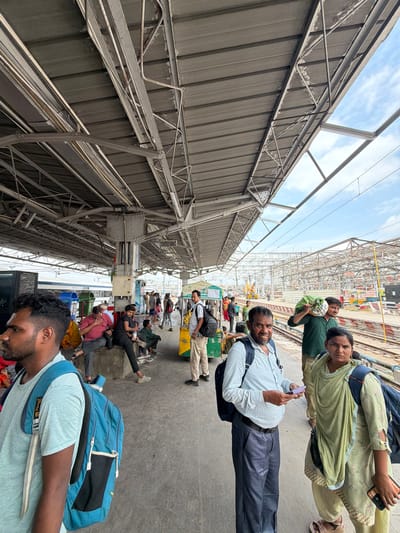 The image depicts a busy platform at a train station in Lucknow, India. A large, covered structure with exposed metal beams and corrugated metal roofing extends over the platform. Several people are present, some sitting on benches or the ground, while others stand or walk. They appear to be waiting for trains or have just arrived. Many are carrying bags and luggage, with a few wearing backpacks.

In the foreground, on the left, a man with dark hair and a beard is seen in profile, wearing a light blue t-shirt and a large blue backpack. To his right, a group of people are seated on a raised concrete ledge. Further down the platform, a man in a light blue patterned shirt and dark pants is holding a small object, possibly a phone or ticket, and looking towards the camera. Beside him, a woman in a light green traditional Indian outfit is also looking towards the camera, holding a mobile phone.

In the background, a train is visible on the tracks. Other individuals and groups of people are scattered throughout the platform. Construction or renovation work appears to be ongoing in the distance, with scaffolding and cranes visible. The overall atmosphere suggests a typical day at an Indian railway station, with people engaged in travel-related activities. The lighting indicates it is daytime, with sunlight filtering through the covered areas. The environment is functional and utilitarian, typical of a public transportation hub.