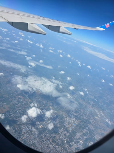 An aerial view from an airplane window shows a sprawling urban landscape beneath a bright blue sky dotted with scattered white clouds. The airplane's wing, painted white with blue accents and a red logo, is visible in the upper portion of the frame, extending towards the right. The land below is a patchwork of agricultural fields and densely populated urban areas, characterized by buildings of various sizes and colors. Roads and waterways crisscross the terrain, connecting different parts of the city. In the distance, an airport with multiple runways is discernible. The overall impression is one of a large, active city seen from a high altitude.
