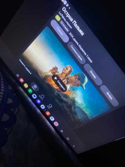 The image shows a close-up view of a tablet screen displaying a streaming service interface. The tablet is held at an angle, suggesting it is being used by someone off-camera. The screen displays the title "Остров Пальма" (Palm Island), information about the series including its rating (6.3), number of reviews (52), genre (drama), country of origin (Norway), and season (1 season). Below the title are buttons for "Главное" (Main), "Сезоны и серии" (Seasons and episodes), "Отзывы" (Reviews), and "Актёры" (Actors). The main visual on the screen is a still from the series, featuring three individuals in the water amidst waves. The tablet has a navigation bar at the bottom with icons for various applications. The overall scene is dimly lit, with the tablet screen being the primary light source. The background is dark and out of focus, with hints of fabric texture, possibly a blanket or upholstery. There is no indication of the tablet's location being in Gergebil, Russia.
