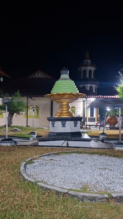 A fountain sculpture is prominently displayed in front of a building that appears to be a mosque at night in Lhokdagang, Indonesia. The fountain's base is a tiered structure with gray and black accents, topped by a large, golden bowl. A green, tiered, conical element sits within the bowl. In the background, a minaret with white and colorful checkered details rises from the mosque. The environment consists of a grassy area with some gravel-filled circular beds and decorative stones. Several lampposts are visible, illuminating the scene. A large, terracotta-colored pot is situated to the right of the fountain. A motorcycle is partially visible under a covered area behind the fountain. The sky is dark, indicating it is nighttime.