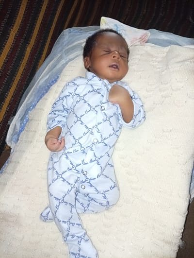 A baby of African descent is sleeping on a white blanket. The baby is wearing a light blue onesie with a repeating "Klain" pattern. The baby has dark hair and closed eyes, and their mouth is slightly open. The blanket has a textured, raised pattern. The baby lies beneath a white and blue mesh netting. The background includes a striped, upholstered surface. There is no information available from this image regarding the specific location in Makera, Nigeria.