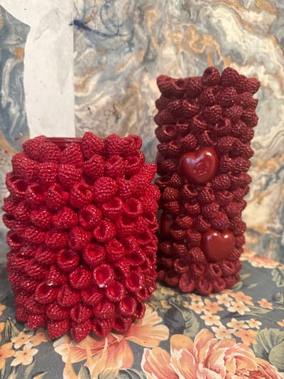 Two red raspberry-shaped candles are displayed on a floral fabric. The candle on the left is shorter and wider, while the candle on the right is taller and thinner. The taller candle has two small red heart-shaped embellishments on its surface. The background consists of a marbled paper with blue, white, and gold tones. The scene is indoors, and there are no people present. The location is not specified in the provided image or context.