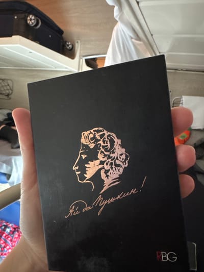 A human hand, with visible fingers and part of the palm, is holding a black rectangular object featuring a copper-toned profile illustration of Alexander Pushkin, facing left. Below the illustration, the Cyrillic text "Ай да Пушкин!" is present in the same copper tone, and the letters "RBG" appear in red and white in the lower right corner. The scene is an interior, specifically located in the city of Gryazi, Russia. In the background, a dark wheeled suitcase rests on a white shelf in the upper left. White fabric is draped in the upper right. A bed or seating area with patterned fabric (red, white, and blue/black) is partially visible in the lower left, indicating a compact accommodation environment, possibly a train compartment.