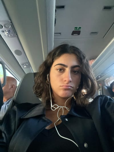 A young female with dark, wavy, shoulder-length hair and brown eyes is centered, looking directly at the camera with a pouting expression. She is seated inside a passenger coach or bus, wearing a black v-neck top and a black collared jacket. White wired earphones are in her ears, with the cable visibly tangled across her chest. To her left, partially visible behind a seat, is the bald head of an older male passenger. The vehicle's interior features grey overhead panels with air vents, reading lights, and an emergency exit sign. This scene occurs within a passenger transport vehicle in the city of Pombal, Portugal.